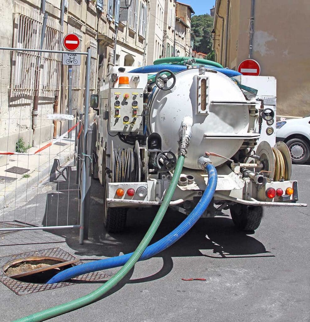 Camion hydrocureur blanc nettoyant un égout via des tuyaux bleus et verts. Scène de voirie urbaine avec panneau sens interdit.