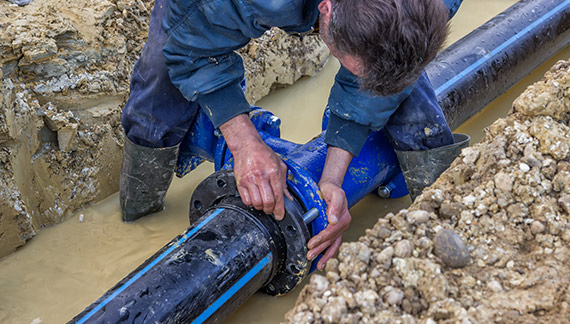 Ouvrier en bottes installe un raccord bleu sur un grand tuyau noir dans une tranchée inondée. Réparation de canalisation.