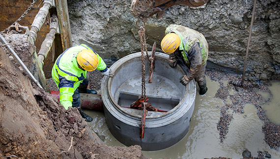 Deux ouvriers en tenue de sécurité posent un élément de tuyau en béton dans une tranchée inondée, avec l'aide d'une grue.