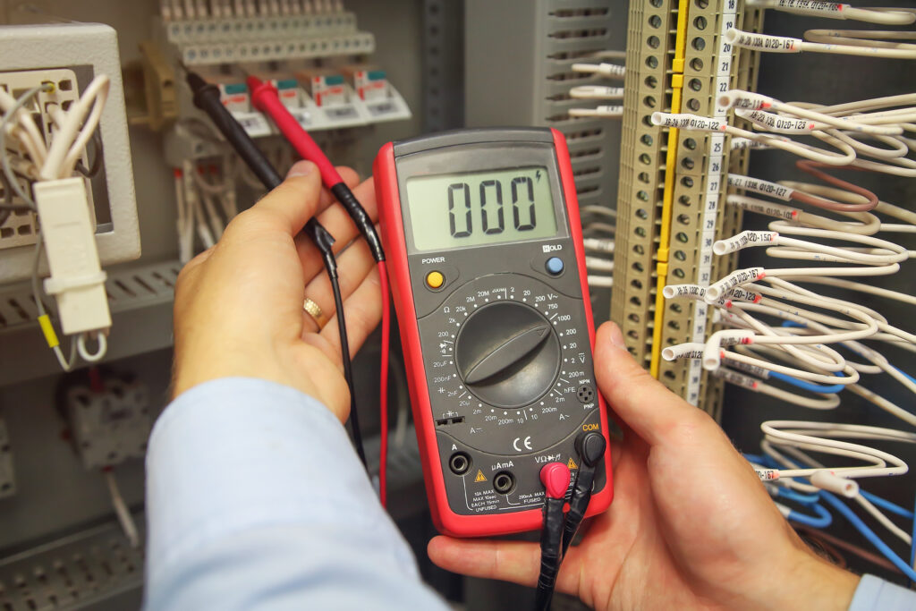 Technicien mesure la tension avec un multimètre numérique rouge affichant '000' dans un tableau électrique. Des fils sont étiquetés.