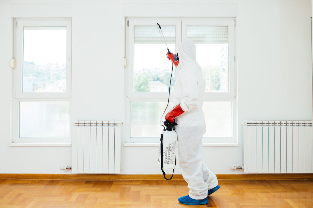 Agent en combinaison de protection blanche et gants rouges pulvérisant avec un 'Pressure Sprayer' dans une pièce vide. Désinsectisation/désinfection.