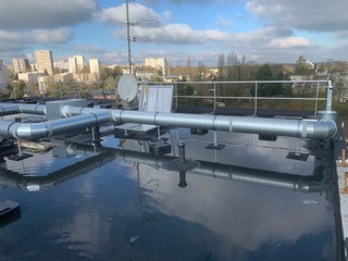 Toiture plate d'immeuble avec de longs conduits de ventilation industriels en acier galvanisé. Garde-corps, antenne et parabole visibles. Ville à l'horizon sous un ciel nuageux.