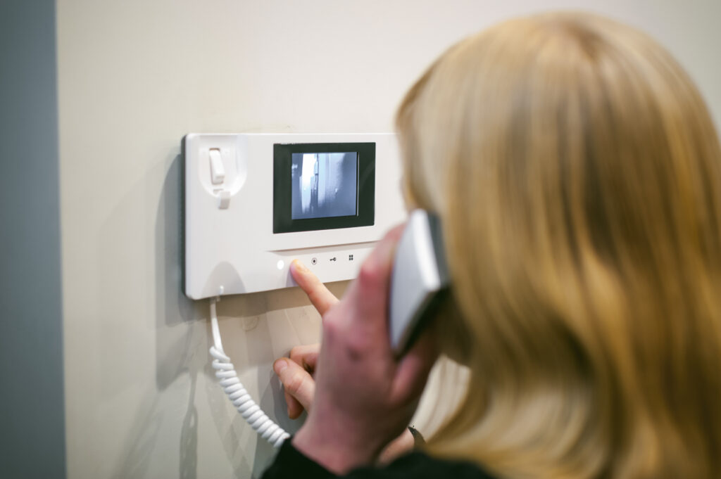 Femme blonde utilisant un interphone mural blanc, appuyant sur un bouton lumineux, écran montrant un couloir. Elle parle au téléphone.