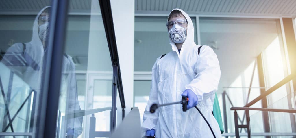 Professionnel en combinaison blanche, masque et lunettes, pulvérisant un produit désinfectant dans un bâtiment moderne et lumineux.