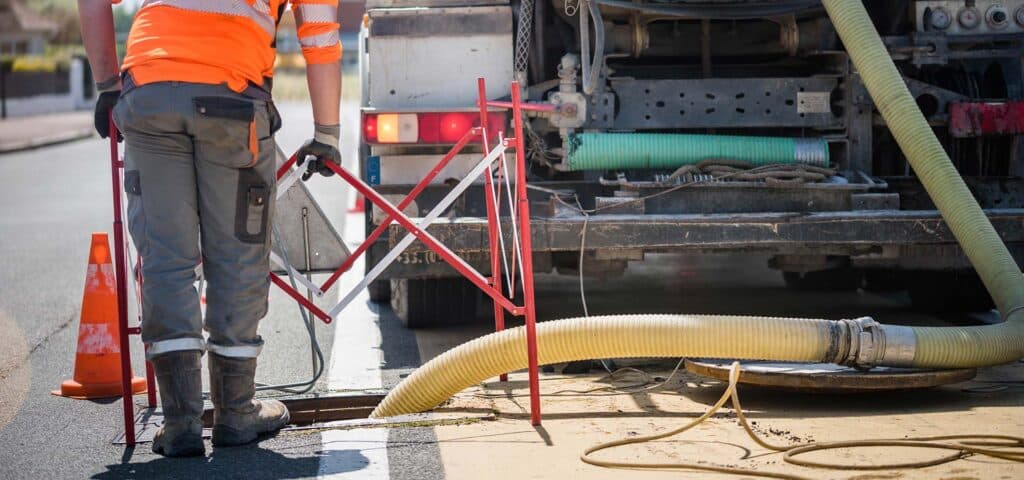 Ouvrier en tenue haute visibilité orange et grise, avec tuyau jaune dans un regard d'égout, près d'un camion et de barrières de chantier.