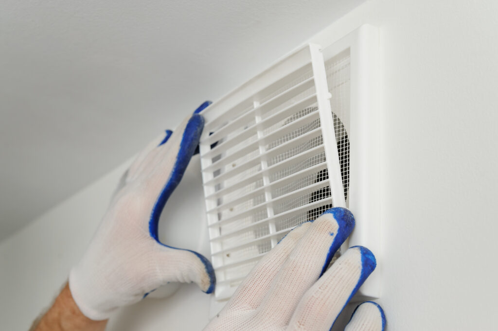 Mains gantées blanches avec bouts bleus installant une grille de ventilation blanche avec moustiquaire sur un mur clair.