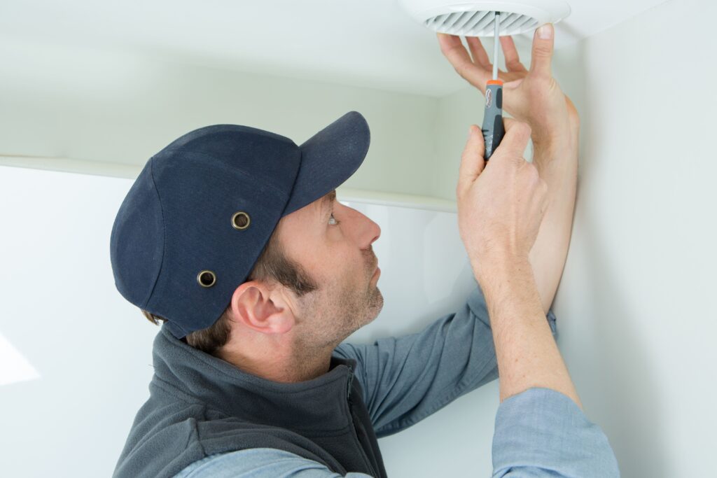 Technicien en casquette bleue et gilet gris fixant une bouche d'aération circulaire au plafond avec un tournevis.