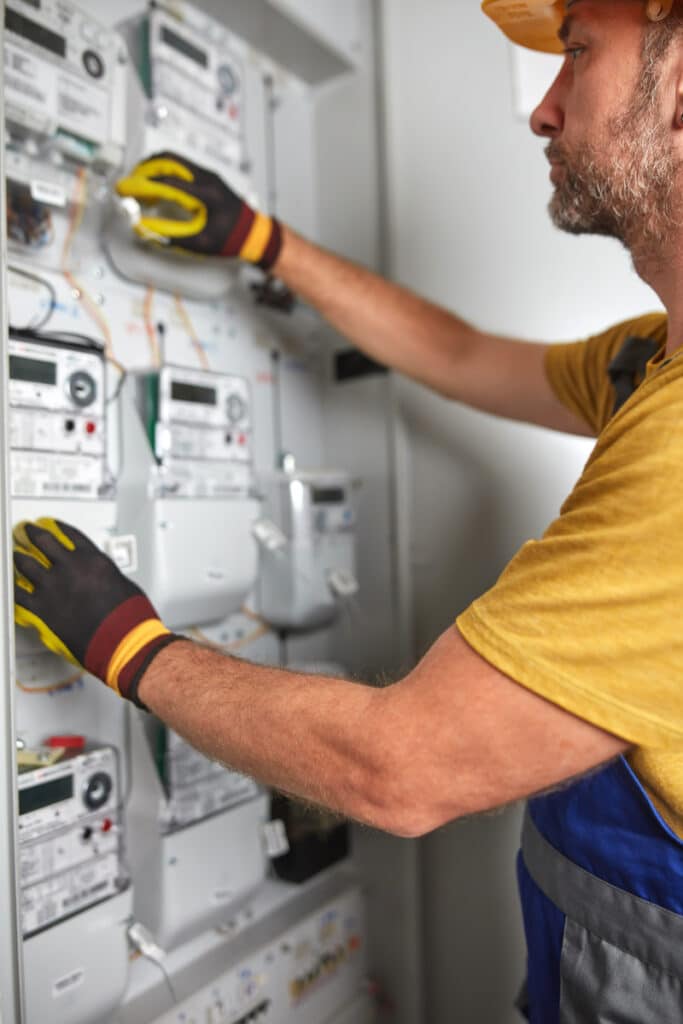Électricien en gants jaunes et casque, ajustant des compteurs électriques sur un tableau de distribution.